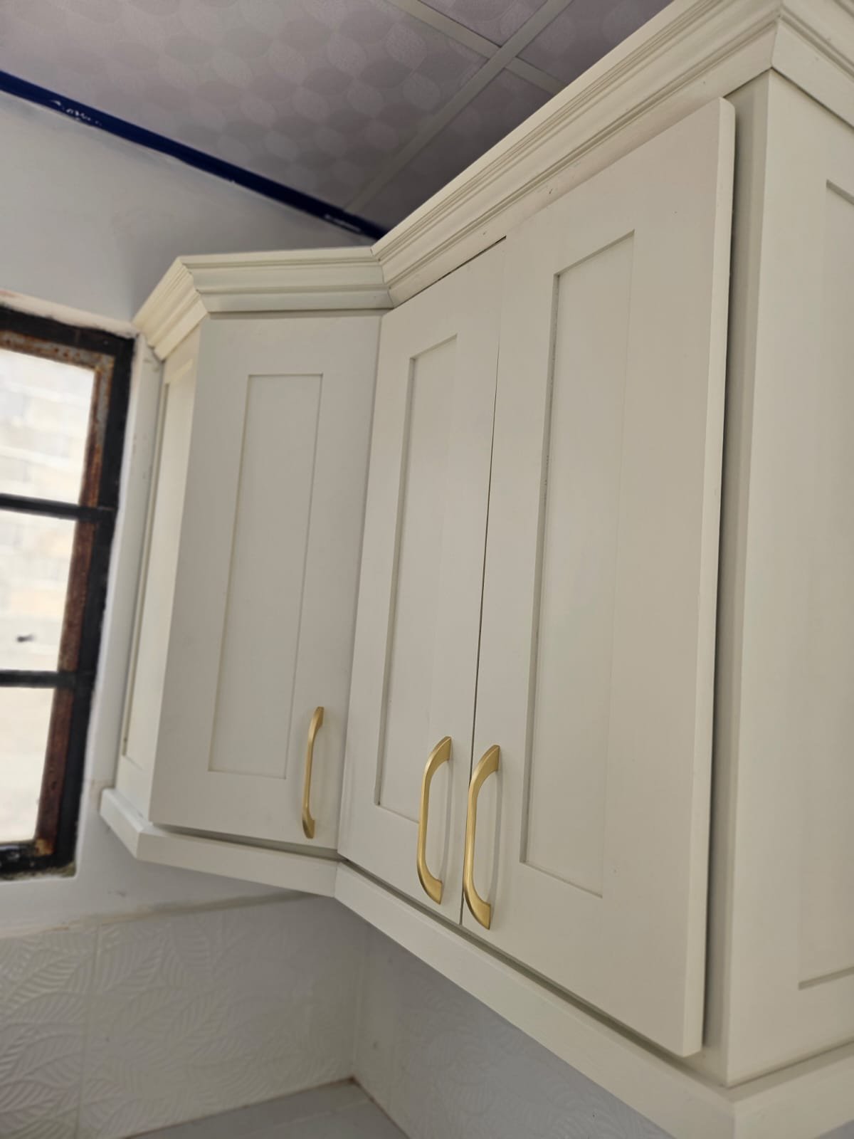 Cream shaker corner wall cabinets with crown moulding beside a kitchen window